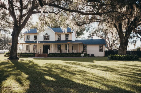 Exterior view of a white two-story farmhouse with a porch, situated on a grassy lawn under large oak trees with Spanish moss.