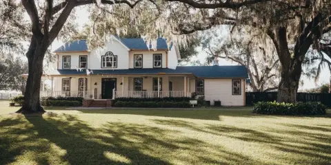 Exterior view of a white two-story farmhouse with a porch, situated on a grassy lawn under large oak trees with Spanish moss.