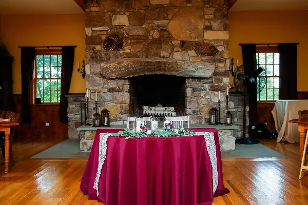 Interior view of a rustic wedding venue featuring a large stone fireplace, hardwood floors, and a head table decorated with a red tablecloth and greenery.