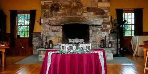 Interior view of a rustic wedding venue featuring a large stone fireplace, hardwood floors, and a head table decorated with a red tablecloth and greenery.