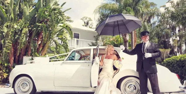 A bride in a white gown stepping out of a classic white car while a chauffeur in a black suit holds a black umbrella over her.