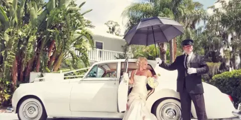 A bride in a white gown stepping out of a classic white car while a chauffeur in a black suit holds a black umbrella over her.