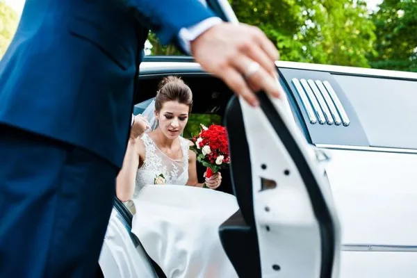 A groom in a blue suit opens the door of a white limousine to help a bride in a white wedding dress and veil exit the vehicle.