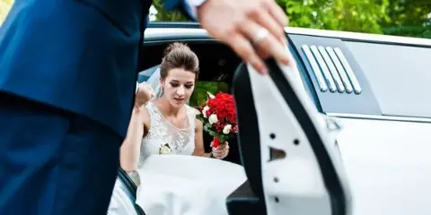A groom in a blue suit opens the door of a white limousine to help a bride in a white wedding dress and veil exit the vehicle.