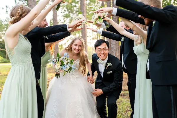 A bride and groom smiling while walking through an arch formed by the hands of their wedding party in a wooded outdoor setting.