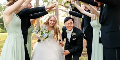 A bride and groom smiling while walking through an arch formed by the hands of their wedding party in a wooded outdoor setting.