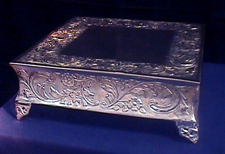 A square silver cake riser with ornate floral detailing on a dark blue surface.