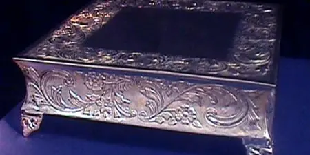 A square silver cake riser with ornate floral detailing on a dark blue surface.
