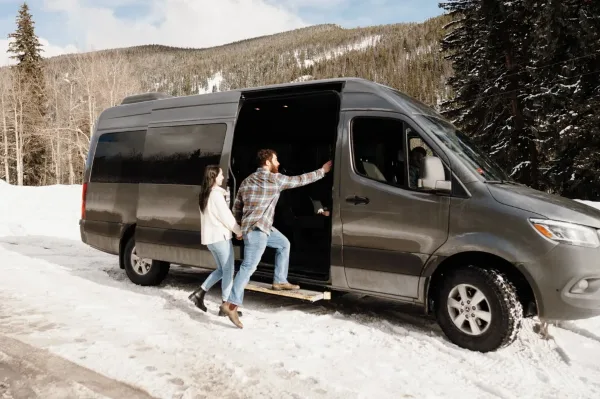 A couple boarding a dark grey luxury Sprinter van in a snowy mountain landscape.