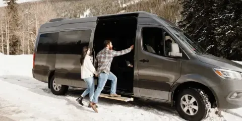 A couple boarding a dark grey luxury Sprinter van in a snowy mountain landscape.