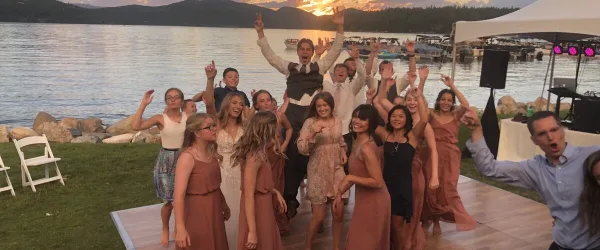 A DJ performing on a stage with professional audio equipment while a group of wedding guests dance at an outdoor lakeside venue.