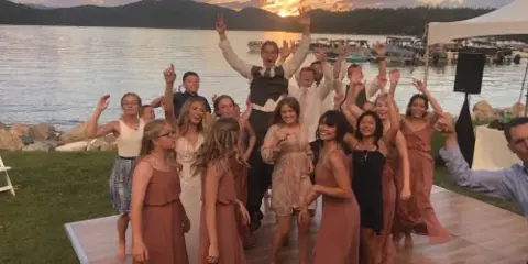 A DJ performing on a stage with professional audio equipment while a group of wedding guests dance at an outdoor lakeside venue.