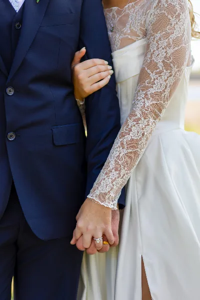 Close-up of a bride and groom holding hands, showing the bride's lace sleeves and wedding ring.
