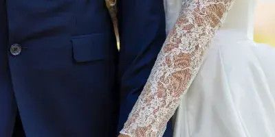 Close-up of a bride and groom holding hands, showing the bride's lace sleeves and wedding ring.