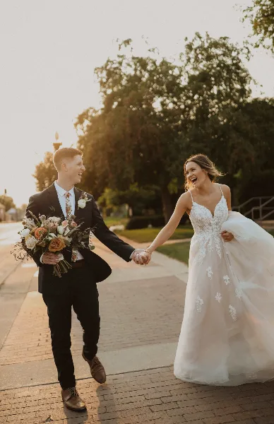 A bride and groom holding hands and laughing while walking on a brick pathway outdoors.