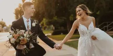 A bride and groom holding hands and laughing while walking on a brick pathway outdoors.