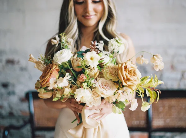 A bride in a white dress holding a large, organic wedding bouquet with cream and peach roses, white ranunculus, and greenery.