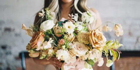 A bride in a white dress holding a large, organic wedding bouquet with cream and peach roses, white ranunculus, and greenery.
