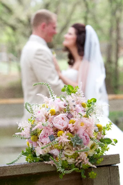 A close-up of a lush bridal bouquet featuring pink peonies, white roses, and greenery, resting on a wooden surface with a blurred wedding couple in the background.