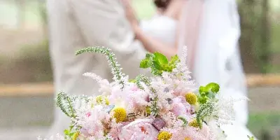 A close-up of a lush bridal bouquet featuring pink peonies, white roses, and greenery, resting on a wooden surface with a blurred wedding couple in the background.