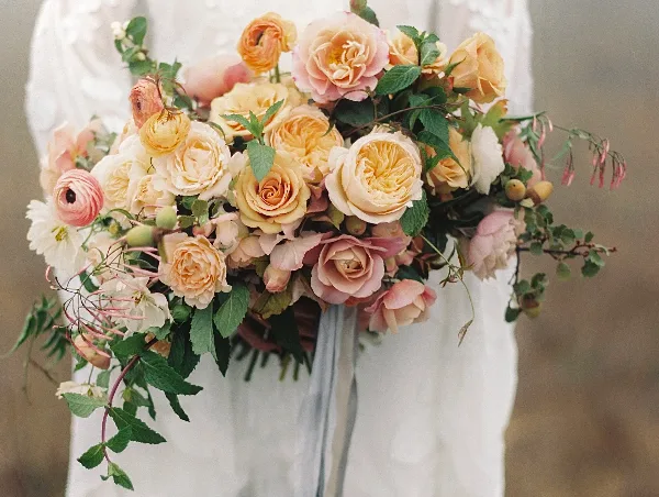 A close-up of a lush bridal bouquet with cream and peach roses, ranunculus, and greenery held by a bride in a white lace dress.