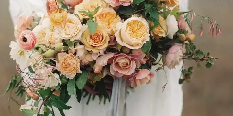 A close-up of a lush bridal bouquet with cream and peach roses, ranunculus, and greenery held by a bride in a white lace dress.