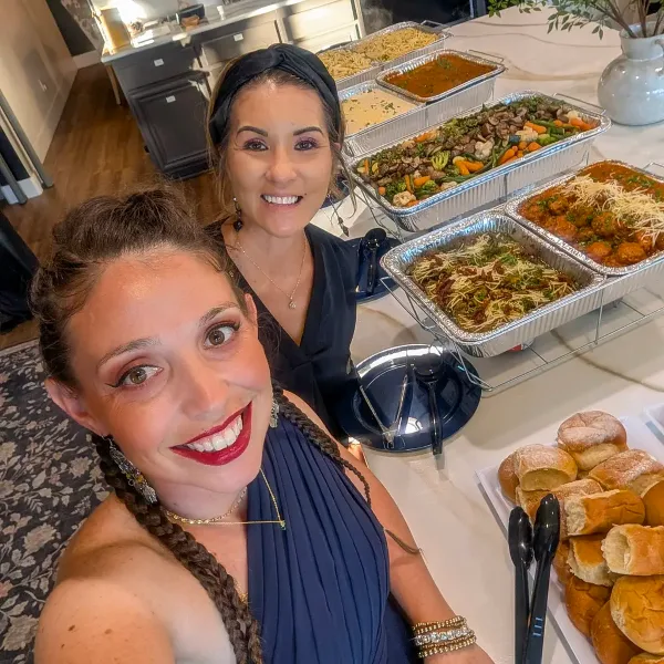 A smiling woman posing with a catering spread of pasta, meatballs, and vegetables in a kitchen.