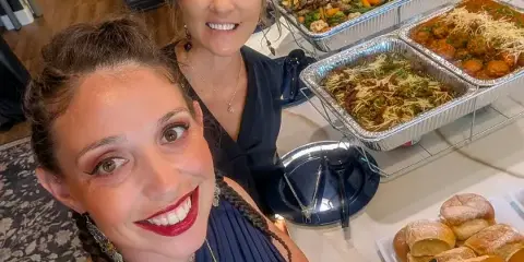A smiling woman posing with a catering spread of pasta, meatballs, and vegetables in a kitchen.