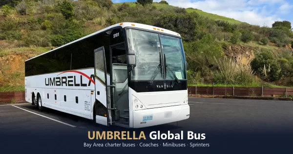 A white Umbrella Global Bus charter coach parked in a lot with a green hillside in the background.