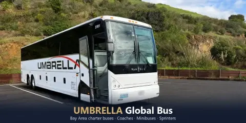 A white Umbrella Global Bus charter coach parked in a lot with a green hillside in the background.