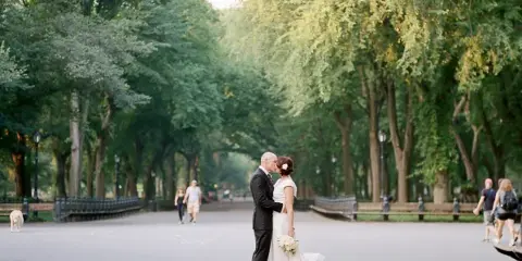 A bride and groom standing in a park with trees in the background.