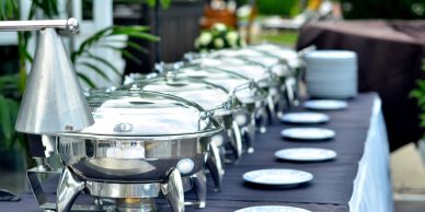 A catering buffet line featuring stainless steel chafing dishes and a row of white plates on a table.