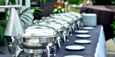 A catering buffet line featuring stainless steel chafing dishes and a row of white plates on a table.