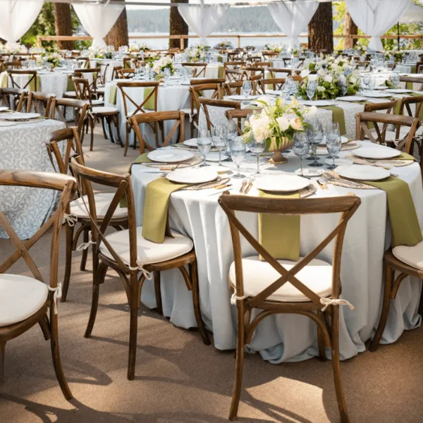 An outdoor event space set with round tables, white tablecloths, green runners, floral centerpieces, and wooden cross-back chairs.