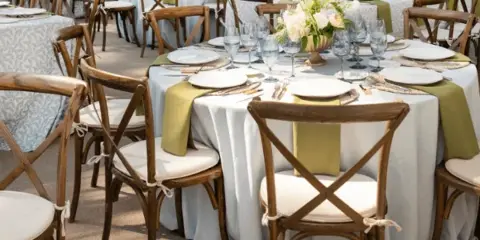 An outdoor event space set with round tables, white tablecloths, green runners, floral centerpieces, and wooden cross-back chairs.