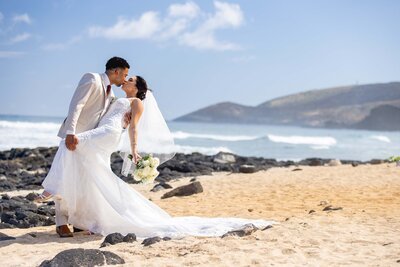 A bride and groom sharing a kiss on a sandy beach in Hawaii during a wedding ceremony.