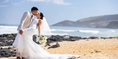 A bride and groom sharing a kiss on a sandy beach in Hawaii during a wedding ceremony.