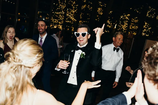 A groom wearing heart-shaped sunglasses dances with guests at a wedding reception.