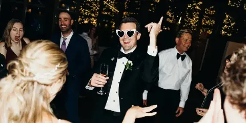 A groom wearing heart-shaped sunglasses dances with guests at a wedding reception.