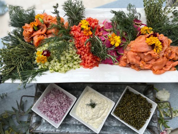 A catering platter featuring smoked salmon, diced tomatoes, cucumbers, and radishes garnished with fresh herbs and edible flowers, accompanied by bowls of capers, onions, and cream cheese.