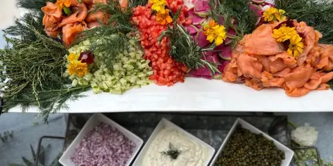 A catering platter featuring smoked salmon, diced tomatoes, cucumbers, and radishes garnished with fresh herbs and edible flowers, accompanied by bowls of capers, onions, and cream cheese.