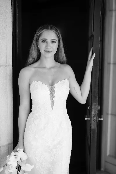 A bride in a white lace gown with styled hair and makeup standing in a doorway.