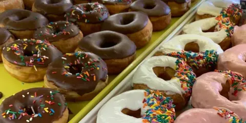 A close-up view of trays filled with chocolate, white, and pink frosted donuts topped with colorful sprinkles.
