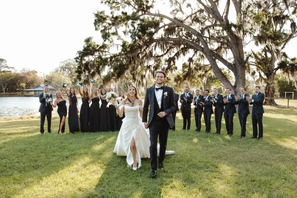 A bride and groom walking down a grassy aisle with their wedding party and a lake in the background.
