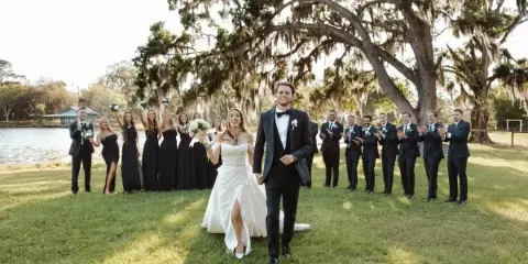 A bride and groom walking down a grassy aisle with their wedding party and a lake in the background.