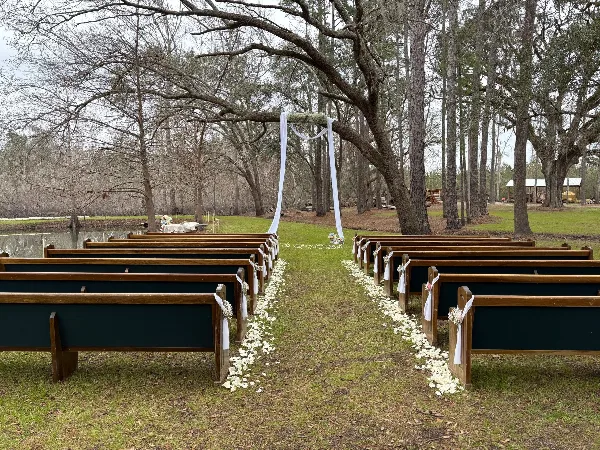 Outdoor wedding ceremony site featuring rows of wooden benches, white floral aisle decorations, and a decorated arch under trees.