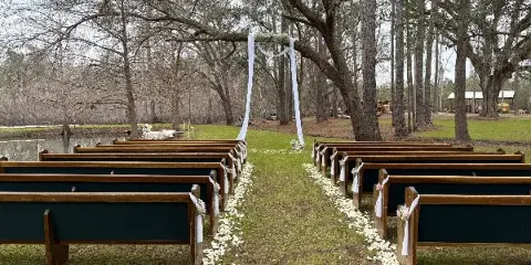 Outdoor wedding ceremony site featuring rows of wooden benches, white floral aisle decorations, and a decorated arch under trees.