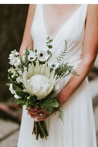 A bride in a white dress holding a large, elegant wedding bouquet featuring a white protea, white anemones, and greenery.