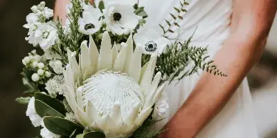 A bride in a white dress holding a large, elegant wedding bouquet featuring a white protea, white anemones, and greenery.