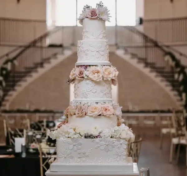A tall, multi-tiered white wedding cake decorated with floral arrangements and fondant flowers, displayed in a large event hall.